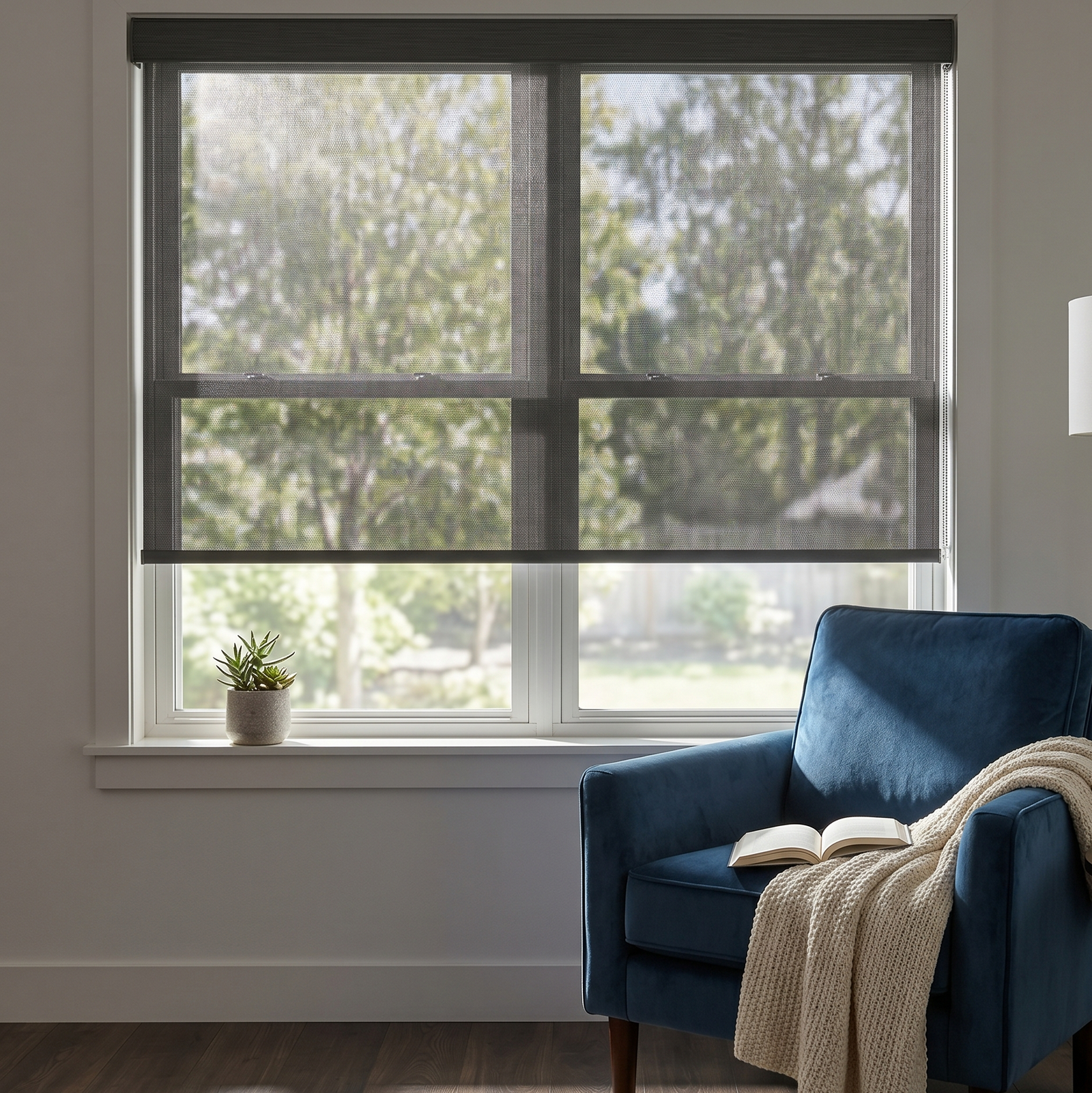 A modern residential living room window featuring a charcoal textured solar screen roller shade partially lowered, filtering bright sunlight. Next to the window is a blue velvet armchair with a book and a knit throw blanket.